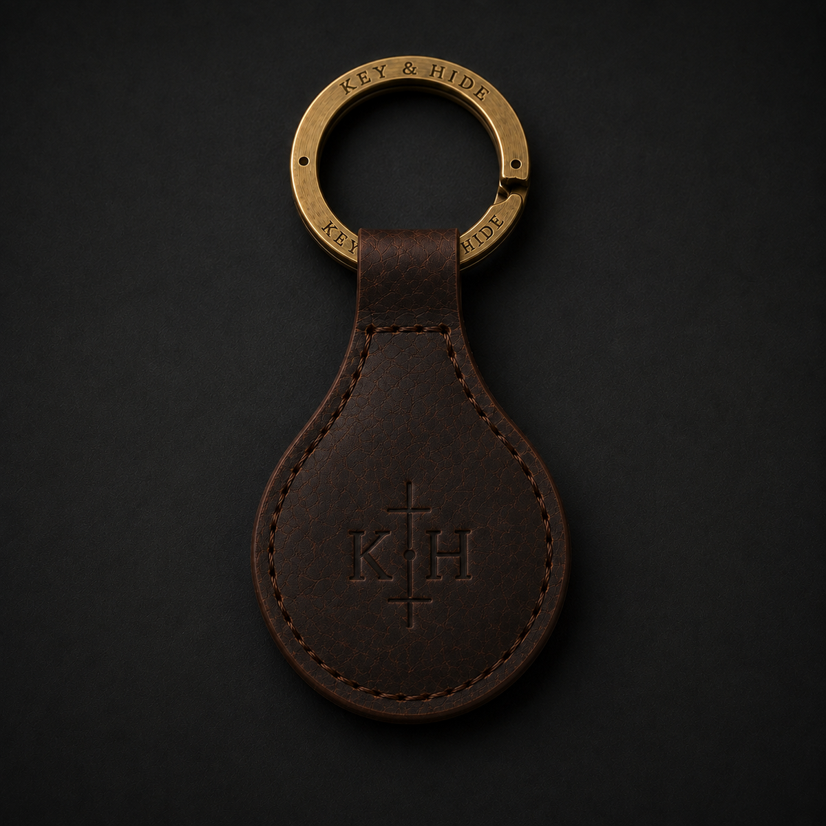 A dark brown leather teardrop keychain with an engraved monogram 'K H' and a brass key ring engraved with 'KEY & HIDE', shown on a dark background.