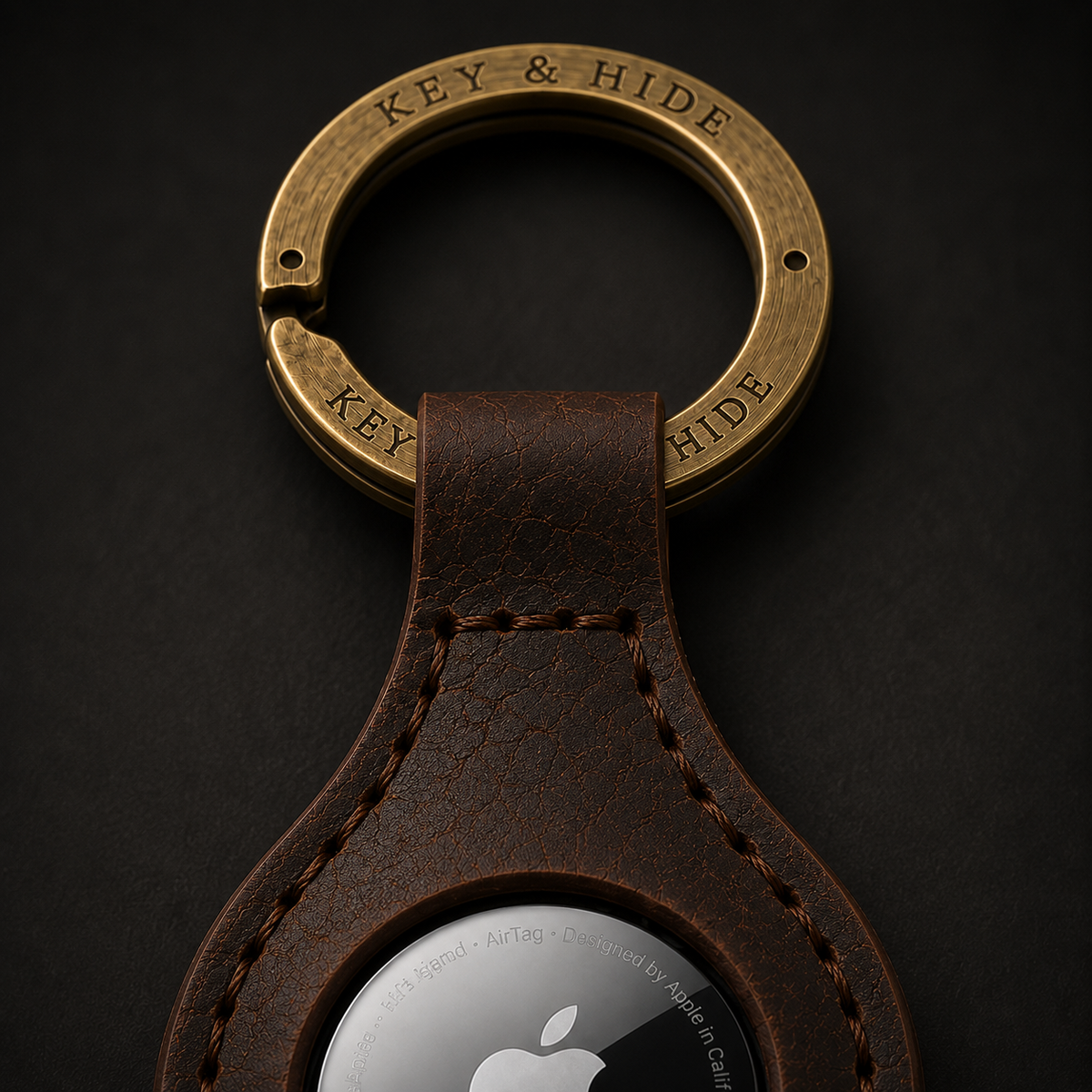 A close-up of a brown leather keychain with a brass key ring engraved with 'KEY & HIDE' and a silver AirTag visible in the leather loop, set against a dark background.