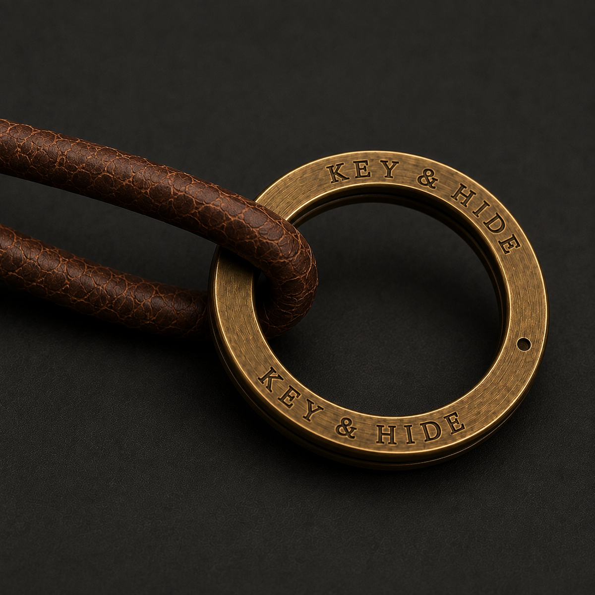 A close-up of a round brass keyring engraved with 'KEY & HIDE' and attached to a brown textured leather cord, set against a dark background.