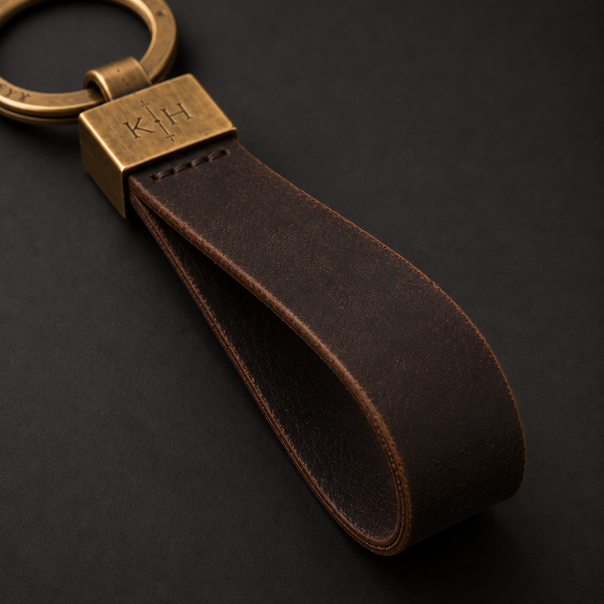 A dark brown leather keychain strap with a brass key ring and a square brass clasp engraved with 'KH', shown on a dark background.