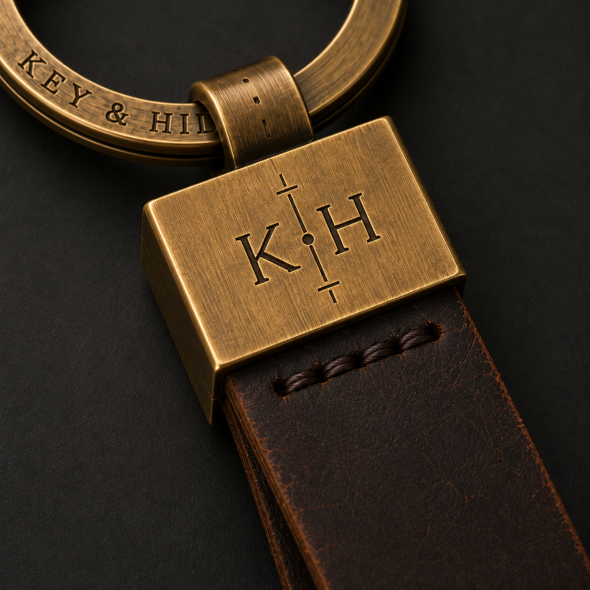 Close-up of a personalized brass keychain with a rectangular engraved plate showing the initials 'K.H' and a dark brown leather strap on a dark background.