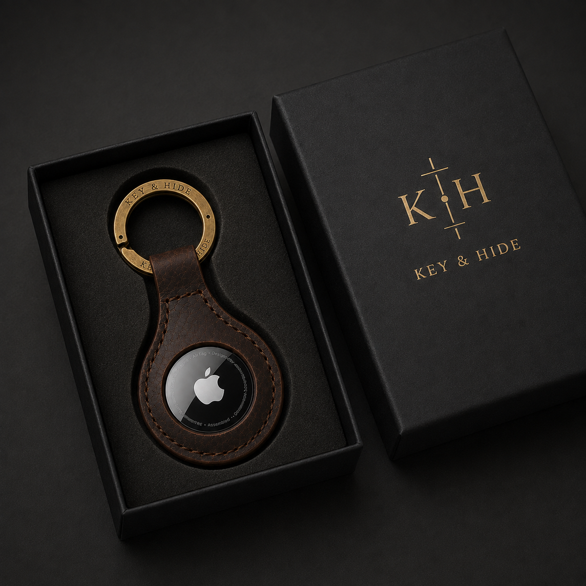 A brown leather keychain with a gold key ring and a black magnetic tracker, presented in a black gift box next to its branded black packaging lid.