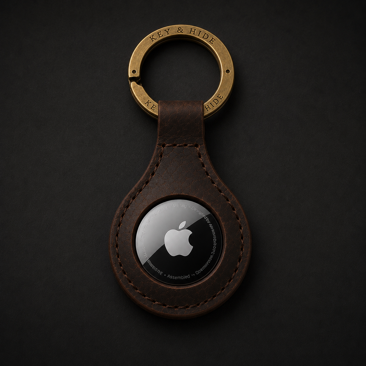 A brown leather keychain with a gold-tone split ring engraved with 'KEY & HIDE' and a black Apple AirTag embedded in the center, shown on a dark textured background.