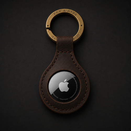 A brown leather keychain with a gold-tone split ring engraved with 'KEY & HIDE' and a black Apple AirTag embedded in the center, shown on a dark textured background.