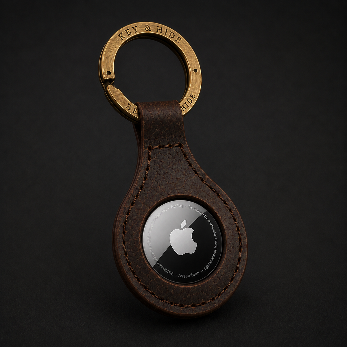 A brown leather key case with a circular cutout containing an Apple AirTag, attached to a brass key ring engraved with 'KEY & HIDE'. The product is photographed on a dark, textured background.